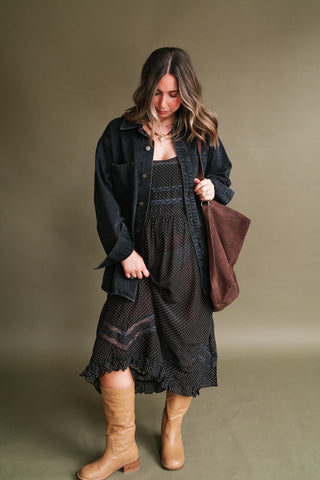Woman wearing a black polka dot lace-trim midi dress layered with a black denim jacket, tan knee-high boots, and a brown shoulder bag.