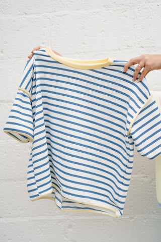 Blue and white horizontal striped relaxed-fit tee featuring a contrast yellow ribbed neckline and trim, held against a white textured brick background.