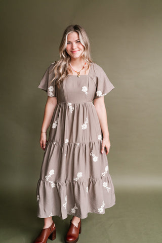 Woman wearing the Evergreen Lane Embroidered Midi Dress in taupe with ivory floral embroidery, a square neckline, flutter sleeves, and tiered skirt, styled with brown heeled shoes and a beaded heart necklace.