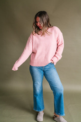 Woman wearing a soft pink knit sweater with a round neckline, center seam detail, long sleeves, and ribbed hem and cuffs, styled with medium-wash straight leg jeans and neutral suede clogs.