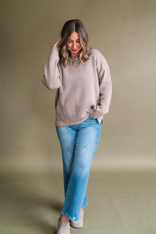 Woman wearing a soft taupe knit sweater with a crew neckline, long sleeves, dropped shoulders, and side slit hem, styled with medium-wash straight leg jeans and neutral suede clogs.