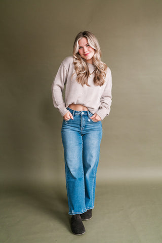 Woman wearing a soft light taupe crewneck sweater with long sleeves and ribbed cuffs, styled with high-rise medium-wash wide leg jeans and dark suede clogs.