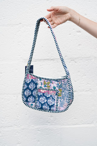 A hand holding a quilted patchwork shoulder bag featuring blue and pink floral patterns with a navy and white striped border against a white textured wall.