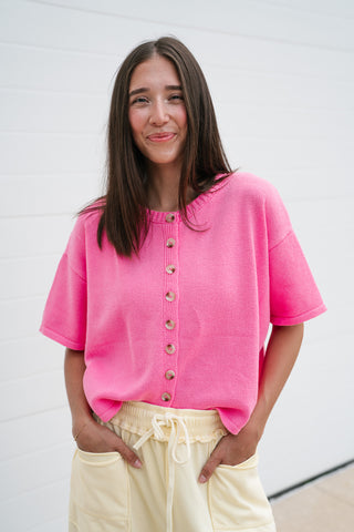 A woman smiling while wearing a vibrant bubblegum pink short-sleeve knit cardigan with brown buttons and light yellow drawstring trousers.