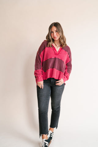 A woman modeling a waffle knit sweater with horizontal stripes in dark red and bright pink, paired with black jeans and black sneakers, standing against a plain beige studio background.
