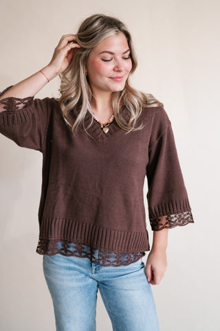 A woman wearing a brown knit sweater with lace trim on the sleeves and hem, paired with light blue jeans, standing against a plain beige background.