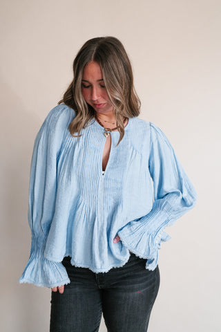 A woman wearing a light blue long-sleeve blouse with a smocked bodice, keyhole neckline, and ruffled cuffs, paired with dark jeans, standing against a plain beige background.