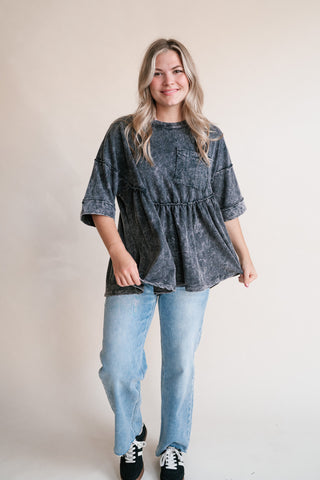 A woman modeling a dark gray acid-wash short-sleeve peplum top with light blue jeans and black sneakers, standing against a plain beige studio background.