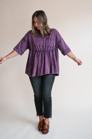 A woman modeling a purple short-sleeve peplum top with a chest pocket, paired with black jeans and brown shoes, against a plain light beige studio background.