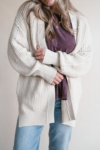 A woman wearing a chunky cream open knit cardigan over a mauve top and light blue jeans, standing against a plain light beige background.
