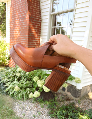A hand holding a brown leather clog-style mule shoe outdoors in front of a house with white siding and a brick column.